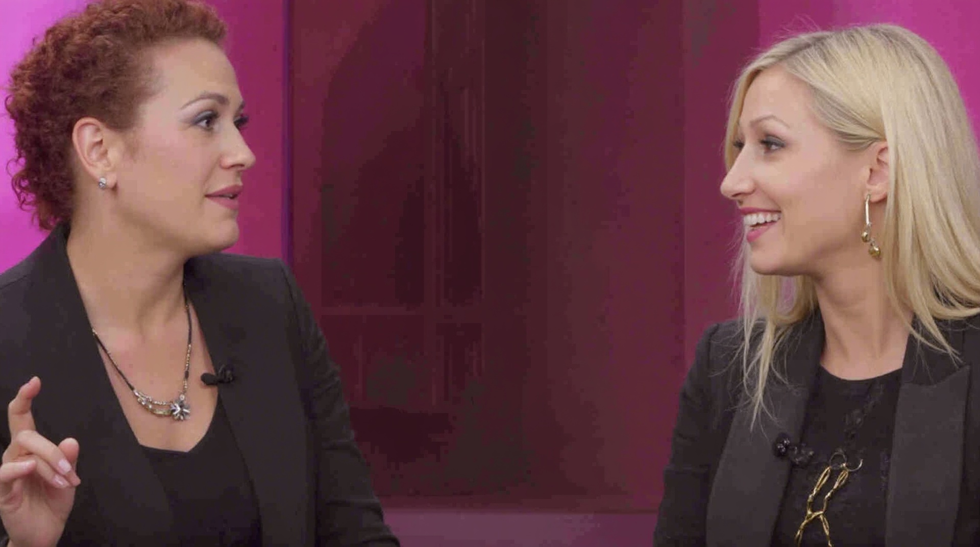 AI generated image by FLUX.1-image-to-image: A YouTube thumbnail featuring two women in conversation against a stylized magenta background with soft lighting and a window silhouette. On the left, a woman with dark red curly hair tied back, wearing a black blazer, earrings, and a necklace, is speaking while raising her index finger with a confident expression. On the right, a blonde woman with light hair styled in a low ponytail, wearing gold hoop earrings and a dark outfit with an embroidered detail, is smiling and looking toward the other woman. The overall mood is professional, engaging, and conversational, resembling an interview or discussion setup.